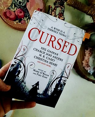 A hand holding a copy of Cursed, edited by Marie O'Regan and Paul Kane