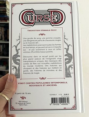 photograph of a man's hand holding a copy of the French edition of Cursed, edited by Marie O'Regan and Paul Kane, to show the back cover