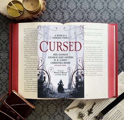 A copy of Cursed, edited by Marie O'Regan and Paul Kane, lying on an open book with sprayed edges that are red, on a wallpapered surface
