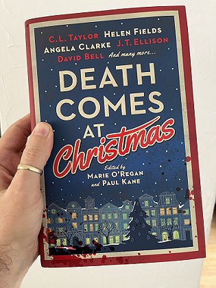 photograph of a man's hand holding up a copy of Death Comes at Christmas, edited by Marie O'Regan and Paul Kane, against a white background