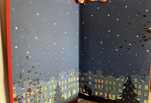 photograph showing the beautiful endpapers for Death Comes at Christmas, edited by Marie O'Regan and Paul Kane. Endpapers feature a row of houses with lit windows, with Christmas trees and garlands in front, and snow falling from a grey sky. Blood is splattered across the left side