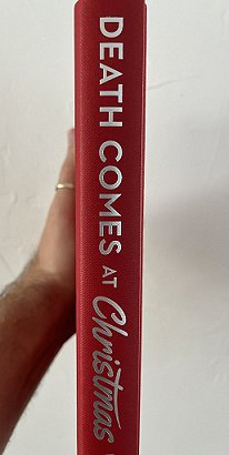 photograph of a man's arm holding up a copy of Death Comes at Christmas, edited by Marie O'Regan and Paul Kane, showing the red bookcover and silver text on the spine