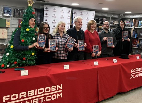 photograph of Death Comes at Christmas editors and authors standing behind a table draped with a red cloth featuring the Forbidden Planet logo, holding copies of Death Comes at Christmas, edited by Marie O'Regan and Paul Kane. L to R: Tina Baker (dressed as a Christmas tree), Fiona Cummins, Helen Fields, Paul Kane, Marie O'Regan, Tom Mead and Russ Thomas (dressed as Death)