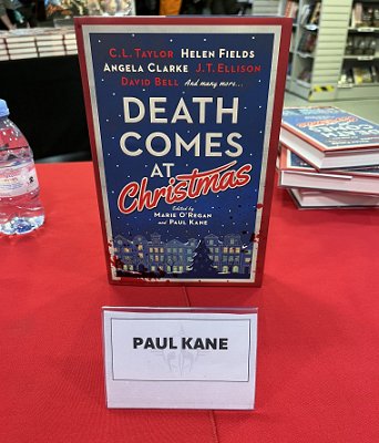 photograph of a copy of Death Comes at Christmas, edited by Marie O'Regan and Paul Kane, standing on a red tablecloth behind a name plate reading Paul Kane. In the background is a bottle of water and three stacked copies of Death Comes at Christmas