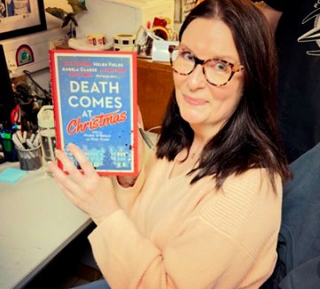 photograph of a smiling Samantha Hayes holding up a copy of Death Comes at Christmas, edited by Marie O'Regan and Paul Kane. In the background is a white desktop with pens, a glass of water and various other items