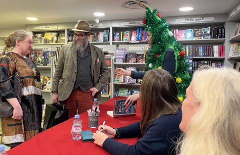 photograph showing Tina Baker chatting to customers wanting their copy of Death Comes at Christmas, edited by Marie O'Regan and Paul Kane, signed. Tina is dressed as a Christmas tree, and in front of her in the photograph are Fiona Cummins and Helen Fields