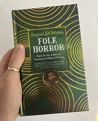 Man's hand holding up a copy of a book, Beyond and Within Folk Horror, short stories edited by Paul Kane and Marie O'Regan