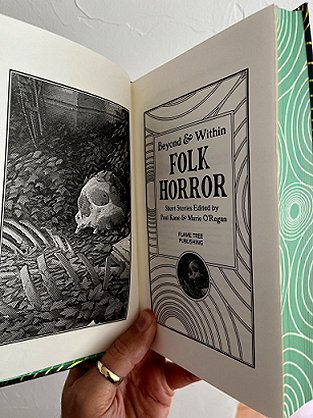 photograph of a man's hand holding up an open copy of Beyond and Within Folk Horror, edited by Paul Kane and Marie O'Regan, to show the title page and accompanying black and white illustration of a skeleton lying on the ground surrounded by fallen leaves