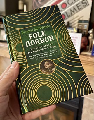 photograph of a man's hand holding up a copy of Beyond and Within Folk Horror, edited by Paul Kane and Marie O'Regan, showing the front cover