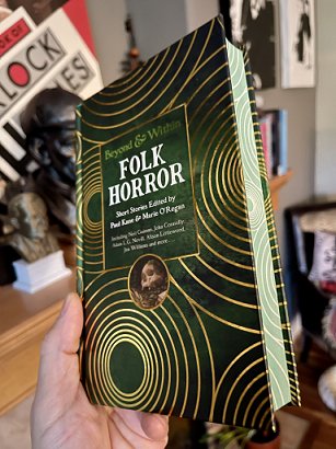 photograph of a man's hand holding up a copy of Beyond and Within Folk Horror, edited by Paul Kane and Marie O'Regan, to show the front cover and sprayed page edges