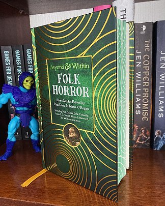 photograph of a copy of Beyond & Within Folk Horror, edited by Paul Kane and Marie O'Regan, on a wooden surface in front of a row of books - a figure of Skeletor stands to the left