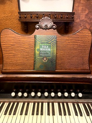 photograph of a copy of Beyond and Within Folk Horror, edited by Paul Kane and Marie O'Regan, standing on the music shelf of a wooden organ