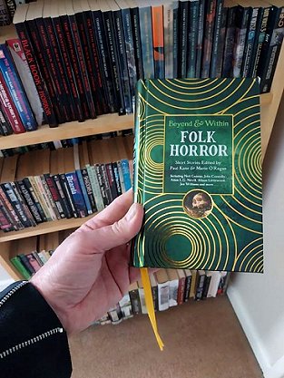 photograph of a man's hand holding up a copy of Beyond and Within Folk Horror, edited by Paul Kane and Marie O'Regan, against a background of a full bookcase