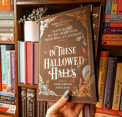 photograph of a woman's hand holding a copy of In These Hallowed Halls, edited by Marie O'Regan and Paul Kane, up aginst a background of bookshelves