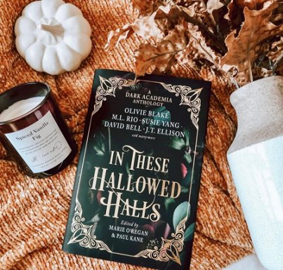 image shows a copy of In These Hallowed Halls, edited by Marie O'Regan and Paul Kane, lying on a rust coloured knitted throw, decorated with a white pumpkin ornament, and candle in a brown glass holder and a white vase full of dried orange leaves