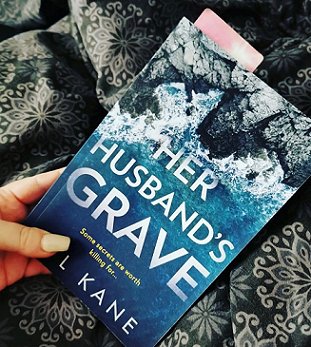 Woman's hand holding a copy of Her Husband's Grave by P L Kane over a patterned cloth