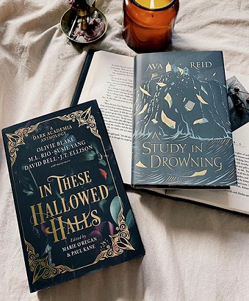 a copy of In These Hallowed Halls, edited by Marie O'Regan and Paul Kane, and a copy of Study in Drowning by Ava Reid, lying on an open book. Beside them are a candle in a jar and a candle holder, on a beige cloth