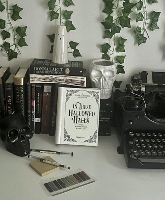 picture showing a copy of In These Hallowed Halls, edited by Marie O'Regan and Paul Kane, open to the title page, standing in front of several books. A black typewriter stands to the righthand side, and ivy climbs the white wall. There are two pens, a paint strip, and black and white skulls on the white desk, and a white ghost statue stands on top of the books at the back