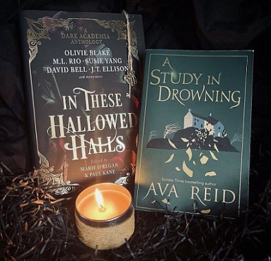 Display featuring a copy of In These Hallowed Halls, edited by Marie O'Regan and Paul Kane, and A Study in Drowning by Ava Reid, with a gold lit candle in front, and standing on black shredded paper