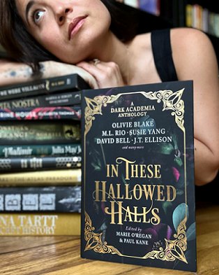 Image of Olivie Blake leaning on a stack of books, behind a standing copy of In These Hallowed Halls, edited by Marie O'Regan and Paul Kane