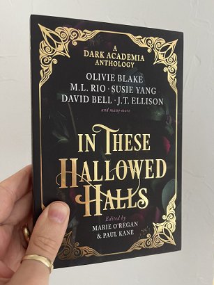 photograph of a man's hand, holding a paperback copy of In These Hallowed Halls, edited by Marie O'Regan and Paul Kane, up against a white background