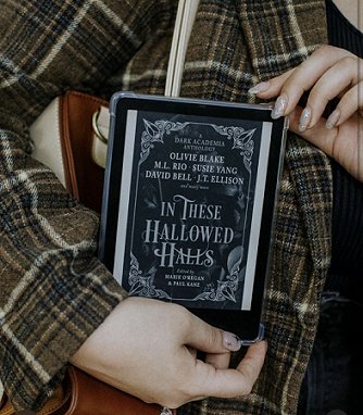 Photograph of a woman in a brown check jacket holding up a tablet with the cover of In These Hallowed Halls, edited by Marie O'Regan and Paul Kane, displayed.