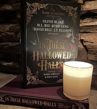Image shows a copy of In These Hallowed Halls, edited by Marie O'Regan and Paul Kane, standing on top of a flat copy of In These Hallowed Halls without a dustjacket, showing the purple cover of the book itself and the gold lettering on the spine. The books are standing in front of brick, and there's a lit votive candle in the forefront of the picture