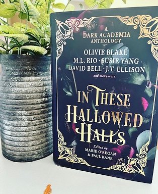 image shows a copy of In These Hallowed Halls, edited by Marie O'Regan and Paul Kane, standing on a white surface, alongside a grey planter with green leaves growing out of the top