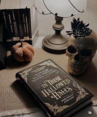 display featuring a copy of In These Hallowed Halls, edited by Marie O'Regan and Paul Kane, lying on a beige woven cloth. Behind the book are a skull filled with black crystals, a cloth pumpkin and a brass lamp, lit with an opaque glass shade, as well as half a dozen standing paperback books with black covers and spines
