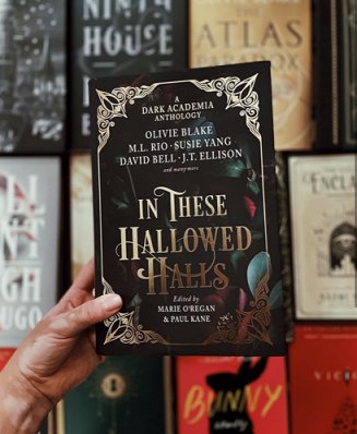 A hand holding a copy of In These Hallowed Halls, edited by Marie O'Regan and Paul Kane, up against a display of Dark Academia novels