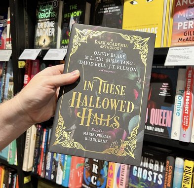 Photograph of a hand holding a copy of In These Hallowed Halls, edited by Marie O'Regan and Paul Kane, in front of rows of bookshelves in a bookstore