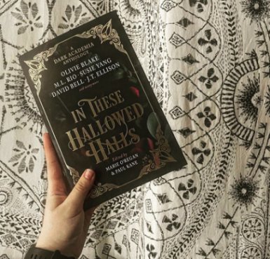 photograph of a hand holding a copy of In These Hallowed Halls, edited by Marie O'Regan and Paul Kane, over a white quilt iwth an intricate black design