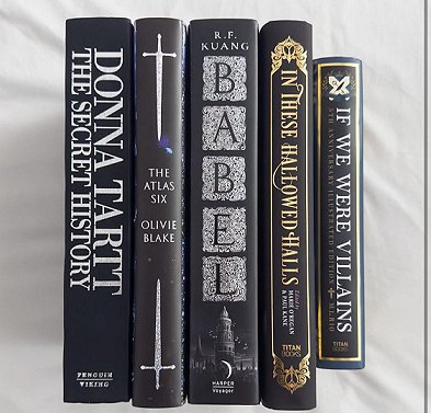 photograph of a display of five books on a white  background, including In These Hallowed Halls, edited by Marie O'Regan and Paul Kane. Other titles are The Secret History by Donna Tartt, The Atlas Six by Olivie Blake, Babel by R F Kuang and If We Were Villains by M L Rio