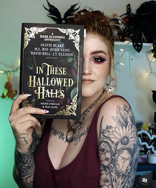 photograph of a young woman in a dark vest top, with tattoos on her arms and chest, holding a copy of In These Hallowed Halls, edited by Marie O'Regan and Paul Kane