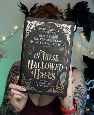 photograph of a young woman with tattoos on her arms, wearing a dark vest top, holding a copy of In These Hallowed Halls, edited by Marie O'Regan and Paul Kane, up in front of her face