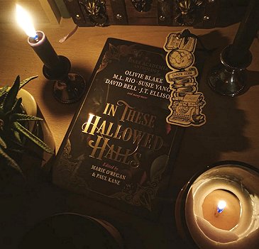 photograph showing a copy of In These Hallowed Halls, edited by Marie O'Regan and Paul Kane, on a wooden surface, lit by candles and with a bookmark on top showing a typewriter, a globe, and a pile of books