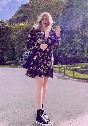 photograph of a woman in a dark floral dress with blonde hair and a black handbag, holding a copy of In These Hallowed Halls, edited by Marie O'Regan and Paul Kane. She's standing on a path with grass and trees in the background