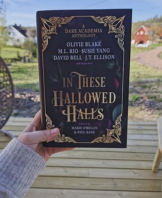 photograph of a woman's hand holding a copy of In These Hallowed Halls, edited by Marie O'Regan and Paul Kane, up against a background featuring a wooden deck, fields and a white and red house with blue sky behind