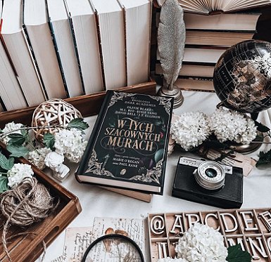 Display featuring a copy of the Polish edition of In These Hallowed Halls, edited by Marie O'Regan and Paul Kane, on a white surface, surrounded by white flowers, wooden letters, a camera and a globe