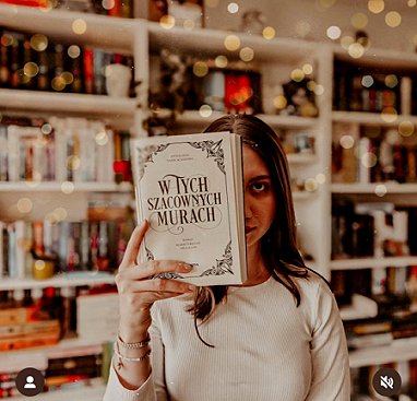 photograph of a woman in a white top holding up the title page of the Polish edition of In These Hallowed Halls, edited by Marie O'Regan and Paul Kane, in front of half her face. Behind here are white bookshelves full of books, lit by fairy lights