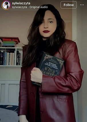 photograph showing a woman with long dark hair and red lipstick, wearing a burgundy leather jacket, holding a copy of the Polish edition of In These Hallowed Halls, edited by Marie O'Regan and Paul Kane. The woman is standing in front of bookshelves and a bed