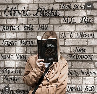 photograph of a woman in a beige jacket holding up a copy of the Polish edition of In These Hallowed Halls, edited by Marie O'Regan and Paul Kane, so you can't see her face, jsut blonde hair. She's standing in front of a brick wall, featuring the names of all the contributors