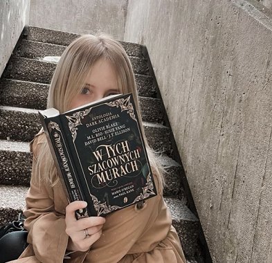 photograph of a young blonde woman sitting on concrete steps, holding an open copy of the Polish edition of In These Hallowed Halls, edited by Marie O'Regan and Paul Kane