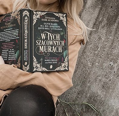 photograph of a young blonde woman sitting against a concrete wall, holding an open copy of In These Hallowed Halls, edited by Marie O'Regan and Paul Kane