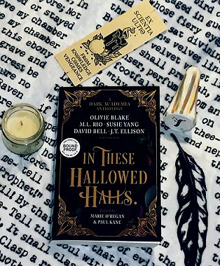 A copy of In These Hallowed Halls, edited by Marie O'Regan and Paul Kane, on a lettered cloth, surrounded by a candle in a glass jar and a bookmark