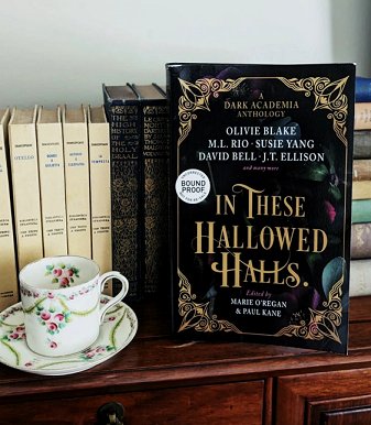 Picture of a standing copy of In These Hallowed Halls, edited by Marie O'Regan and Paul Kane, in front of  row of books and beside a teacup and saucer