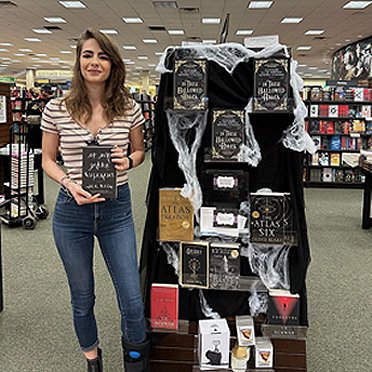 image showing M L Rio holding a copy of If We Were Villains, beside a book display on a black background laced with cobwebs, featuring In These Hallowed Halls, edited by Marie O'Regan and Paul Kane