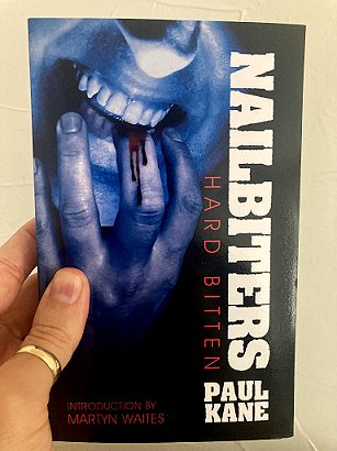 image of a man's hand with a gold ring on the thumb holding a copy of a book with a blue-tinted man's face, the man is biting fingers hard enough to make them bleed. Title Nailbiters Hard Bitten by Paul Kane
