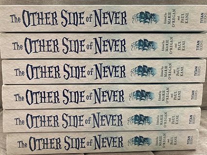 Stack of six copies of The Other Side of Never, edited by Marie O'Regan and Paul Kane, showing the book spines with the title and the image of a sailing ship