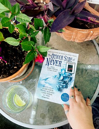 A woman's hand holding a copy of The Other Side of Never, edited by Marie O'Regan and Paul Kane, on a glass table surrounded by pot plants and a glass with clear liquid and a slice of lemon
